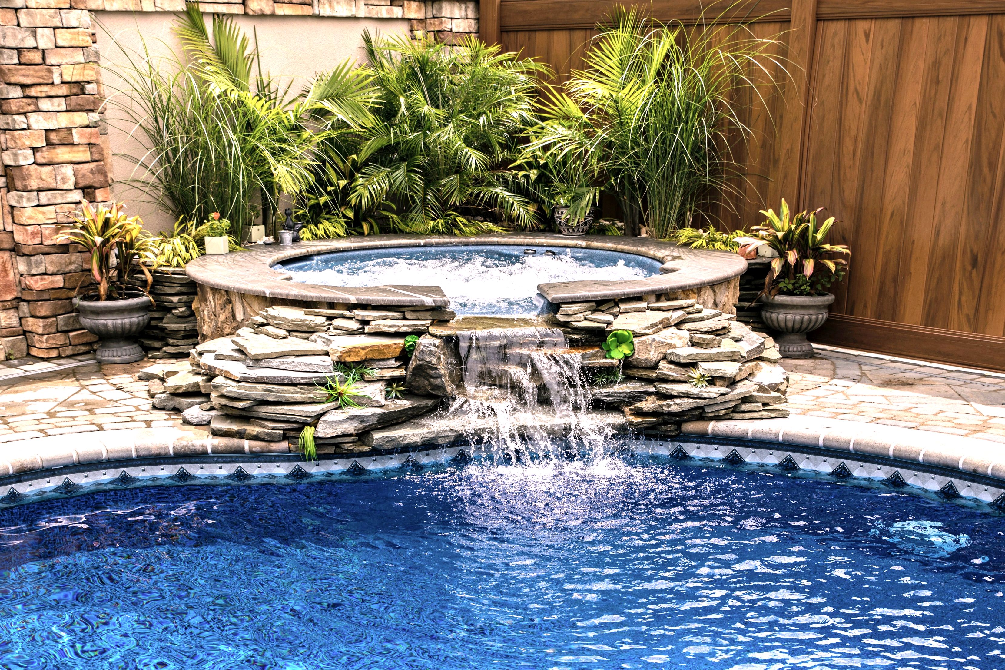 Stone spillover spa with waterfall, palms and wood fence behind pool